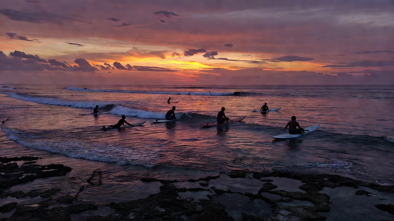 Surfers silhouetted over a reef lineup at dusk in Indonesia